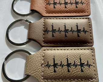 May include: Three leather keychains with silver key rings. Each keychain has a brown, tan, or gray leather strap with a black silhouette of a rifle and a heartbeat graphic. The text on the keychain reads "My Heart Beats for Freedom".