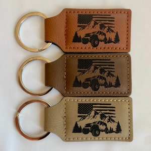 May include: Three leather keychains with a silver key ring. Each keychain features a brown leather rectangle with an engraved design of a jeep driving through a mountain landscape with an American flag in the background. The keychains are brown, dark brown, and light brown.
