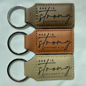 Può includere: Tre portachiavi in pelle marrone e beige. Ogni portachiavi ha un anello argentato e un'etichetta rettangolare con le parole "SHE IS STRONG" e "PROVERBS 31:25" stampate in nero.