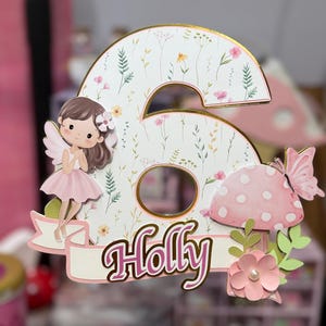 May include: A whimsical birthday cake topper in the shape of the number six, featuring a fairy, mushroom, and butterfly. The number is decorated with floral patterns and the name "Holly" in pink script.