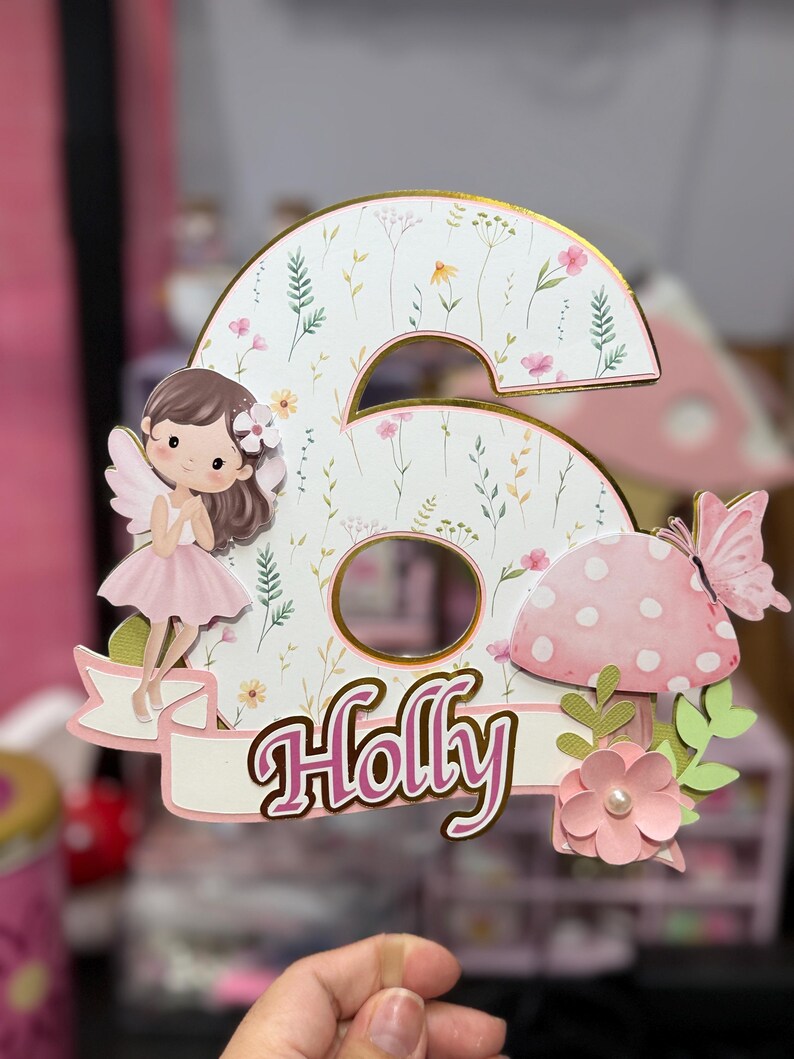 May include: A whimsical birthday cake topper in the shape of the number six, featuring a fairy, mushroom, and butterfly. The number is decorated with floral patterns and the name "Holly" in pink script.
