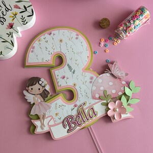 May include: A pink and green layered number three cake topper with a floral pattern. It features a fairy, a mushroom, and the name "Bella" on a banner. The topper is designed for a birthday celebration.