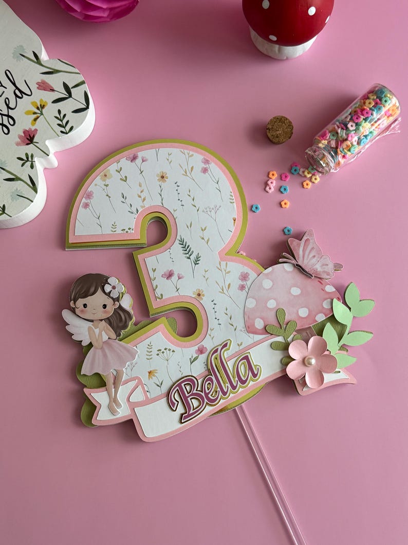 May include: A pink and green layered number three cake topper with a floral pattern. It features a fairy, a mushroom, and the name "Bella" on a banner. The topper is designed for a birthday celebration.