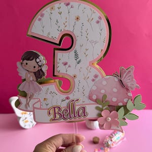 May include: A whimsical cake topper featuring a large number 3 with a floral pattern, gold trim, and the name "Bella." It includes a fairy, mushroom, and butterfly accents, perfect for a birthday celebration.