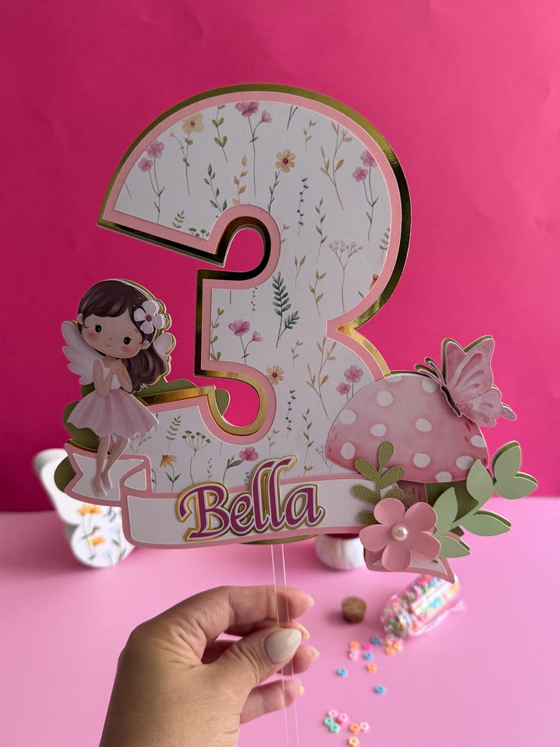 May include: A whimsical cake topper featuring a large number 3 with a floral pattern, gold trim, and the name "Bella." It includes a fairy, mushroom, and butterfly accents, perfect for a birthday celebration.