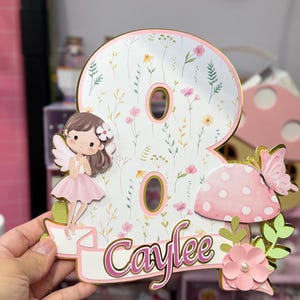 May include: A whimsical birthday cake topper in the shape of the number eight, featuring a fairy, mushroom, and butterfly. The number is decorated with a floral pattern and the name "Caylee" in pink script. The topper has gold accents.