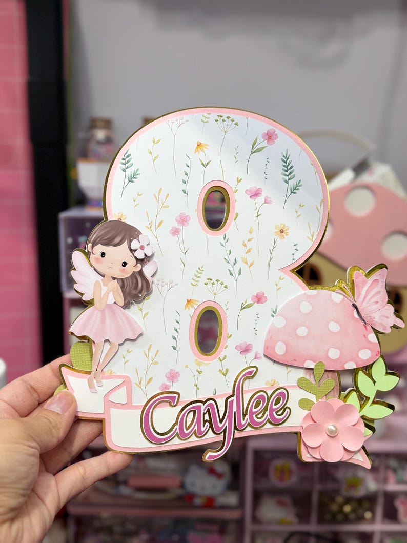 May include: A whimsical birthday cake topper in the shape of the number eight, featuring a fairy, mushroom, and butterfly. The number is decorated with a floral pattern and the name "Caylee" in pink script. The topper has gold accents.