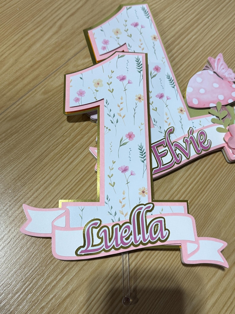 May include: A pink and gold number one cake topper with floral accents and the name "Luella" in cursive script. The number is decorated with a floral pattern and the name "Elvie" is also visible.