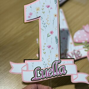 May include: A floral-patterned number one cake topper with a gold border and pink accents. The name "Luella" is displayed on a banner below the number. The design includes pink and yellow flowers and green leaves.