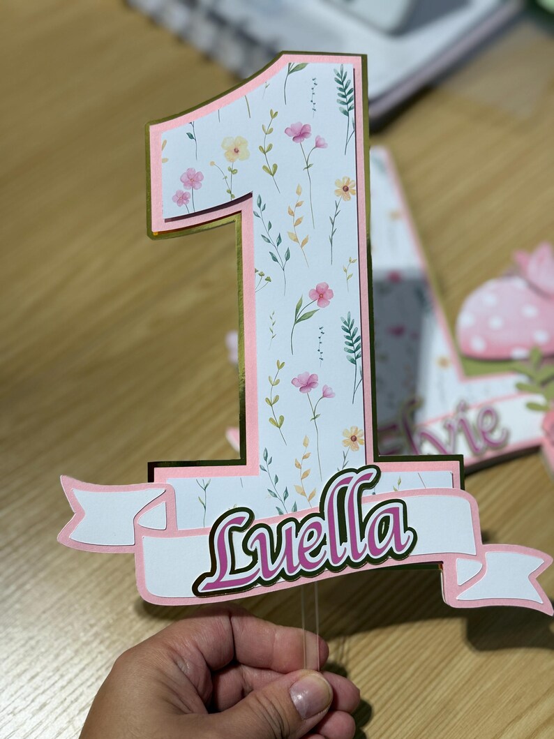 May include: A floral-patterned number one cake topper with a gold border and pink accents. The name "Luella" is displayed on a banner below the number. The design includes pink and yellow flowers and green leaves.
