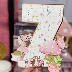 May include: A decorative number seven cake topper with a floral design and the name "Ellie". The topper features a fairy, a pink mushroom, and a butterfly. The number is decorated with a gold border.