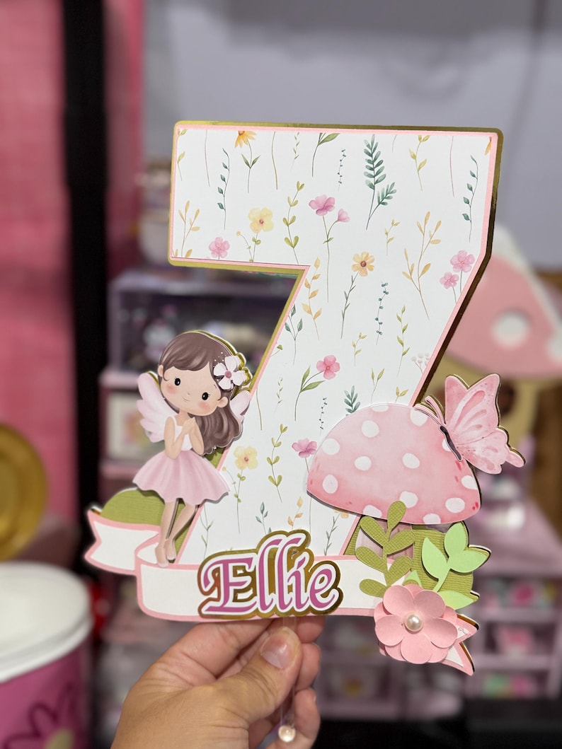 May include: A decorative number seven cake topper with a floral design and the name "Ellie". The topper features a fairy, a pink mushroom, and a butterfly. The number is decorated with a gold border.