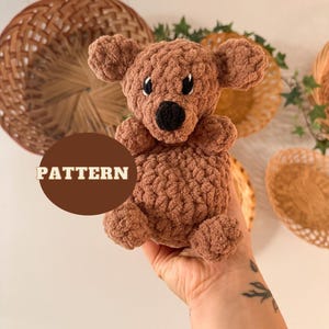 May include: A brown crocheted stuffed animal shaped like a dog. The dog has black eyes and a black nose. The word "PATTERN" is written in yellow text on a brown circle.