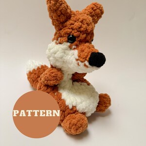 May include: A crocheted fox plush toy with orange and white fur. The fox is sitting with its tail curled behind it. The toy has black eyes and a black nose. The word "PATTERN" is written in white letters on a brown circle.