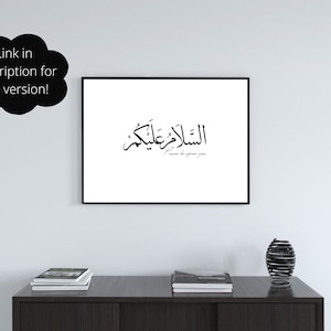 May include: A black and white framed print with Arabic calligraphy that reads "The peace be upon you" in English. The print is hanging on a white wall above a dark brown table with two stacks of books and a black metal decorative vase.