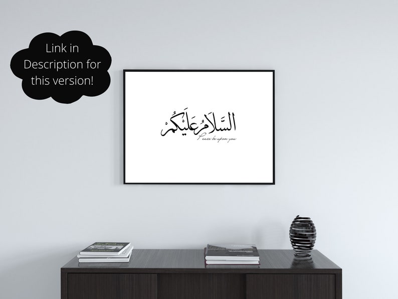 May include: A black and white framed print with Arabic calligraphy that reads "The peace be upon you" in English. The print is hanging on a white wall above a dark brown table with two stacks of books and a black metal decorative vase.