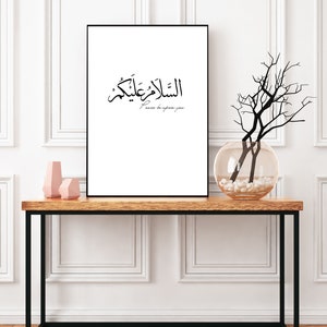 May include: A black and white print with Arabic calligraphy that reads "السلآم عليك" which translates to "Peace be upon you". The print is framed and hanging on a white wall above a wooden console table with a vase of branches and two pink ceramic vases.