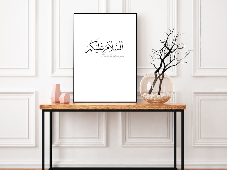 May include: A black and white print with Arabic calligraphy that reads "السلآم عليك" which translates to "Peace be upon you". The print is framed and hanging on a white wall above a wooden console table with a vase of branches and two pink ceramic vases.