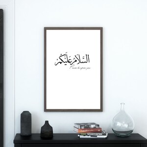 May include: A framed print with Arabic calligraphy that reads "Peace be upon you" in English. The print is hanging on a white wall in a modern interior.