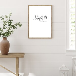 May include: A framed print with the Arabic phrase "Al Salam Alaykum" and the English translation "Peace be upon you". The print is hanging on a white wall with a wooden plank background.