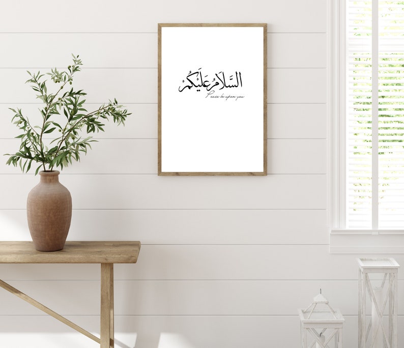 May include: A framed print with the Arabic phrase "Al Salam Alaykum" and the English translation "Peace be upon you". The print is hanging on a white wall with a wooden plank background.