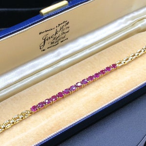 Könnte beinhalten: Ein goldenes Armband mit einer Reihe von rubinroten Edelsteinen, die in einer goldenen Kette eingefasst sind. Das Armband befindet sich in einer blauen Samt-Schmuckschatulle mit weißer Satin-Auskleidung. Die Schachtel trägt ein Etikett mit der Aufschrift "Jacks of London, 78 New Bond Street, London W1."