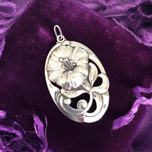 May include: A silver pendant featuring a detailed floral design with a delicate flower and intricate scrollwork. The pendant is oval-shaped and has a polished finish.