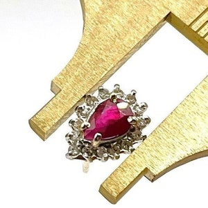 May include: A close-up of a ruby ring with a pear-shaped ruby surrounded by a halo of smaller diamonds. The ring is being measured with a ruler.