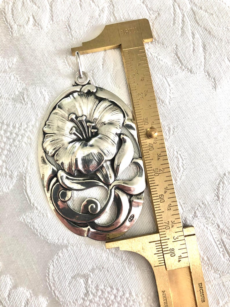 May include: A silver pendant featuring a detailed floral design with a single flower and leaves. The pendant is approximately 5 cm in length and has a loop at the top for attaching a chain.