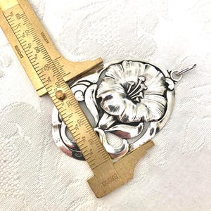 May include: A silver pendant featuring a detailed floral design with a large flower in the centre. The pendant is approximately 5 cm in length.