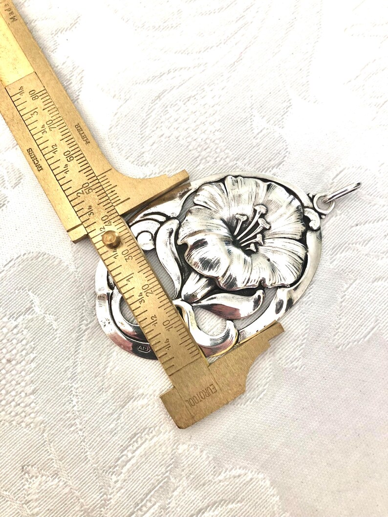 May include: A silver pendant featuring a detailed floral design with a large flower in the centre. The pendant is approximately 5 cm in length.
