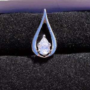 Könnte beinhalten: Ein silberner Anhänger in Tropfenform mit einem birnenförmigen, klaren Edelstein in der Mitte. Der Anhänger ist vor einem schwarzen, strukturierten Hintergrund platziert, der den Glanz und das Design des Schmucks hervorhebt.