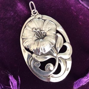 May include: A silver pendant featuring a detailed, stylised flower design with a delicate, openwork pattern. The pendant is oval-shaped and has a loop at the top for attaching a chain.