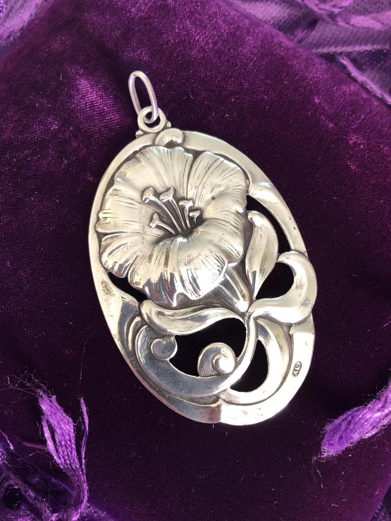 May include: A silver pendant featuring a detailed, stylised flower design with a delicate, openwork pattern. The pendant is oval-shaped and has a loop at the top for attaching a chain.