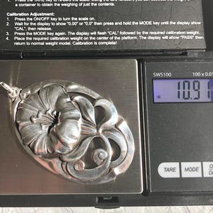 May include: A silver pendant shaped like a flower with intricate details, weighing 10.9 grams on a digital scale.