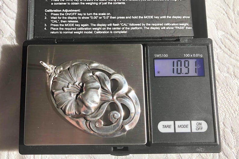 May include: A silver pendant shaped like a flower with intricate details, weighing 10.9 grams on a digital scale.