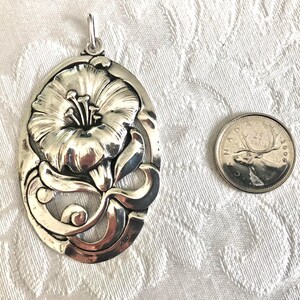May include: A silver pendant featuring a detailed, stylised flower design. The pendant is oval-shaped and has a loop at the top for attaching a chain.