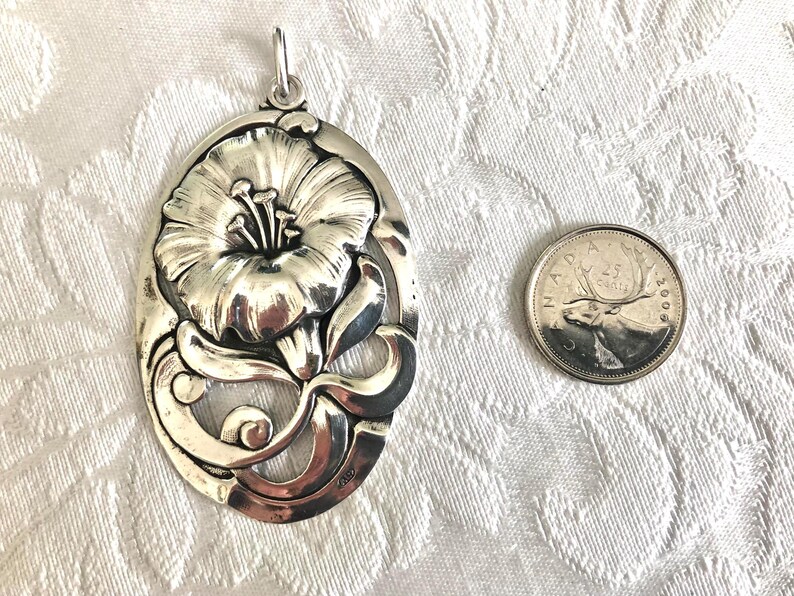 May include: A silver pendant featuring a detailed, stylised flower design. The pendant is oval-shaped and has a loop at the top for attaching a chain.