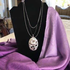 May include: A silver necklace with a floral pendant on a black display stand. The necklace is layered with three chains and the pendant is a detailed oval shape with a flower design. The stand is draped with a purple scarf.