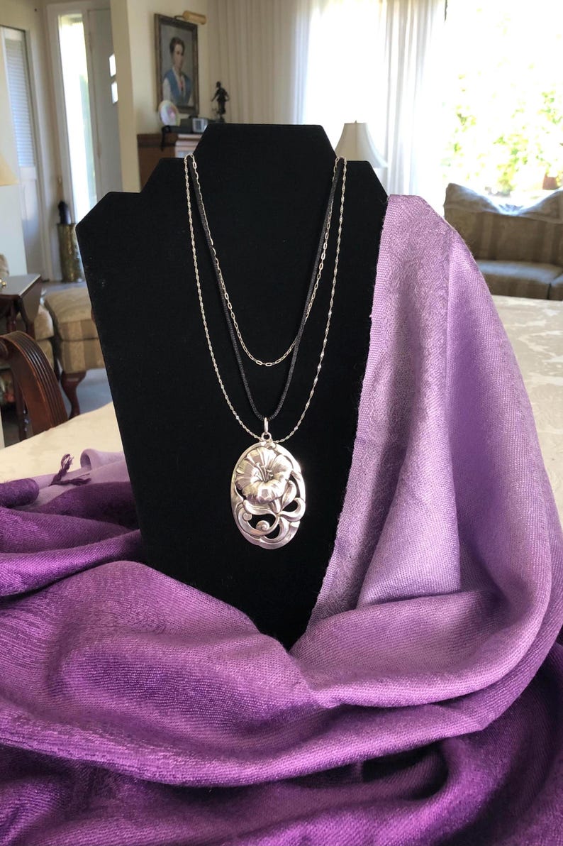 May include: A silver necklace with a floral pendant on a black display stand. The necklace is layered with three chains and the pendant is a detailed oval shape with a flower design. The stand is draped with a purple scarf.