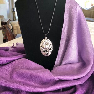May include: A silver pendant necklace with a floral design hangs on a black display stand. The pendant is oval shaped and features a detailed flower with leaves. The necklace is displayed on a purple and white ombre scarf.