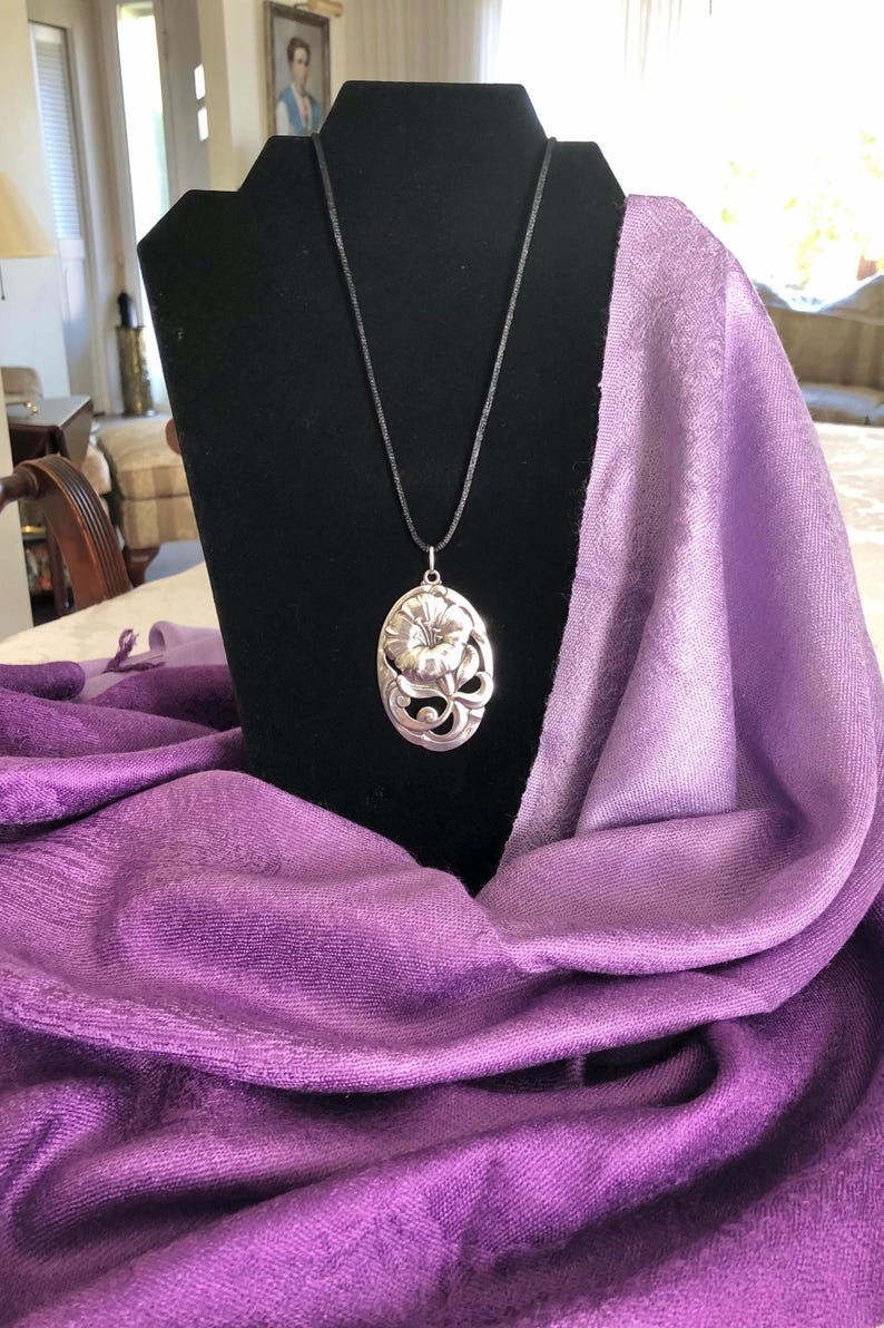 May include: A silver pendant necklace with a floral design hangs on a black display stand. The pendant is oval shaped and features a detailed flower with leaves. The necklace is displayed on a purple and white ombre scarf.