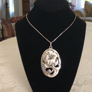 May include: A silver necklace with a large, detailed flower pendant. The pendant is oval-shaped and features a stylised flower with intricate details.