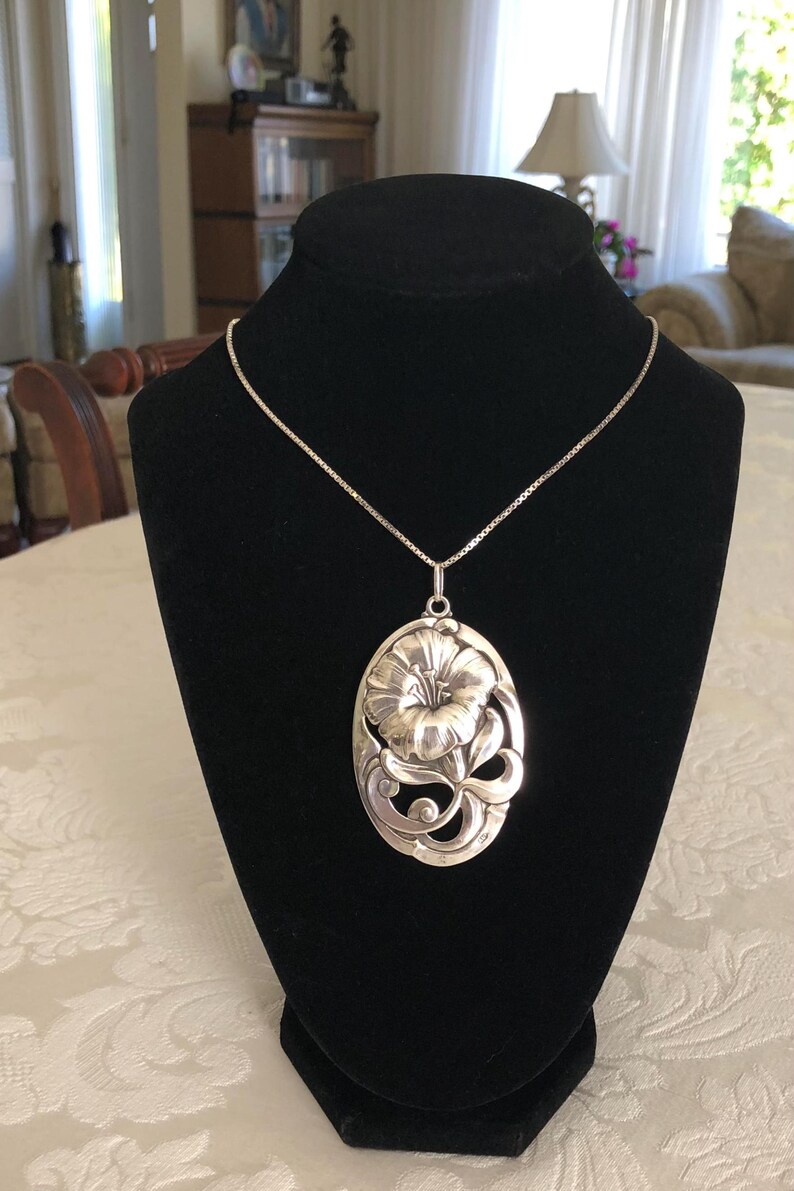 May include: A silver necklace with a large, detailed flower pendant. The pendant is oval-shaped and features a stylised flower with intricate details.