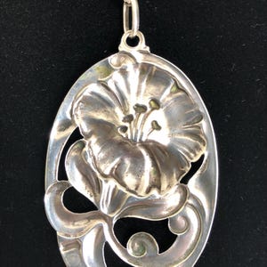 May include: A silver pendant necklace featuring a detailed floral design. The pendant is oval-shaped and has a raised flower with intricate details. The flower is surrounded by a delicate, swirling design.
