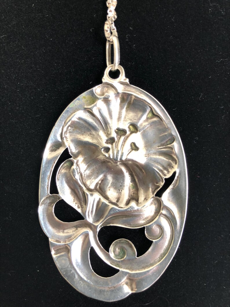 May include: A silver pendant necklace featuring a detailed floral design. The pendant is oval-shaped and has a raised flower with intricate details. The flower is surrounded by a delicate, swirling design.