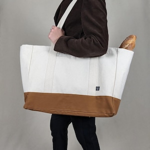 May include: A large canvas tote bag with a white upper section and a brown base. The bag has long, cream-coloured straps and a small logo patch. A loaf of bread is visible in the bag. The bag is being carried by a person.