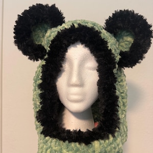 May include: A light green crochet balaclava featuring a thick, fuzzy black trim around the face opening and two large, round, fuzzy black ears attached to the top. The textured yarn has a soft, plush appearance, creating a distinct bear-like look.