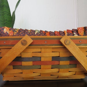 May include: A rectangular woven basket with wooden handles and a decorative fabric trim featuring autumn leaves. The basket is made of interwoven strips in shades of orange, green, and brown, with a dark brown trim and wooden handles.