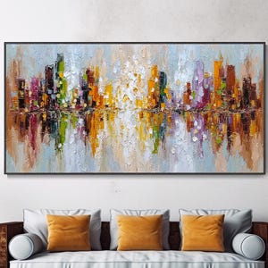 May include: An abstract cityscape painting in a black frame. The artwork features a skyline with colorful buildings in shades of orange, yellow, green, purple, and brown, reflected in a mirrored effect below. The painting hangs above a brown sofa with orange pillows.